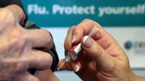 A close up of a nurse's hand with French manicured nails holding a syringe poised to vaccinate a patient's shoulder. A blurred sign on the wall behind says Flu, Protect Yourself.