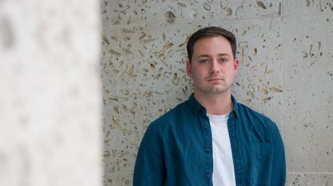 Ben is a 25-year-old man and stood against a pale white stone background outside. He is in a blue shirt with a white t-shirt underneath, has brown hair combed to the side and has one hand in his pocket