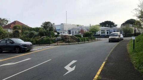 The image shows a two-lane road approaching what appears to be a school or public building, with cars travelling in both directions and a narrow pavement running along the right-hand side.
