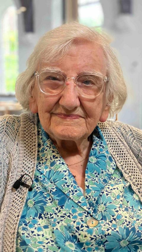 Marjorie, an elderly woman, looks at the camera with a small smile. She has short white hair, and wears clear glasses, a blue floral print shirt and a light cardigan. 