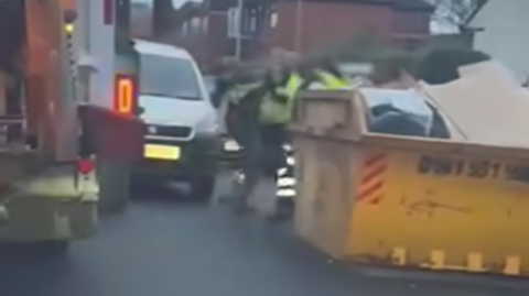 A number of men in reflective jackets grapple with each other, partially obscured by a yellow waste skip. On the left of the frame is the back of a bin lorry, and a white van is visible just behind the men. 