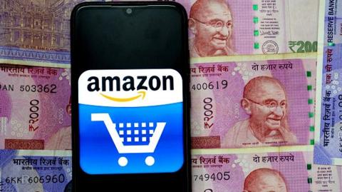 In this photo illustration an Amazon logo seen displayed on a smartphone with the Indian Currency Note One Hundred and Two thousand on the background.