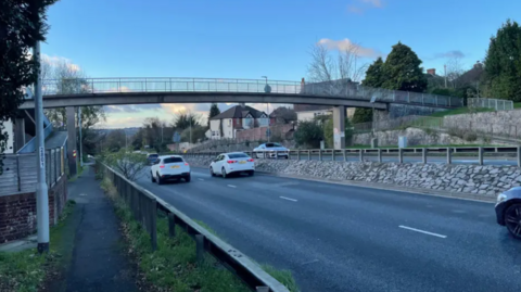 A footbridge crossing over a highway with multiple cars pictured on the road, with houses to the right of it and a blue sky in the background. 