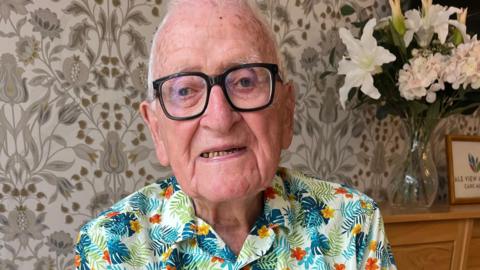 Dennis, who has short grey hair, black-rimmed glasses and a Hawaiian shirt, is looking at the camera and smiling. Some lilies are in a vase on a sideboard in the background, to the right.