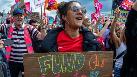 A large group of teachers and education staff on strike are gathered in a street raising their hands and holding flags and placards. In the foreground, a woman with dark hair tied back, hoop earrings and sunglasses, holds a sign that says "fund our schools". She wears a red t-shirt under a black coat. Behind her, a man wearing a blue hat, striped top and a high-vis pink vest holds a flag.
