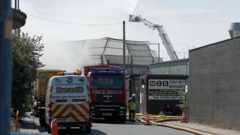 A fire appliance parked on a road in front of an industrial unit, with hoese running along the street. A cherry picker is visible behind. 