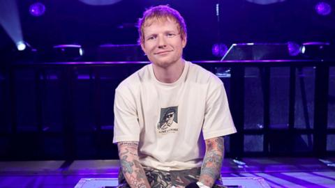 Ed Sheeran wearing a white T-shirt. He's in a room lit by purple light.