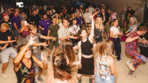 A group of people dancing underneath a marquee. It is dark outside and there are strings of lights draped across the top of the marquee.