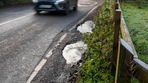 A road travels along a country road. Some rain-filled potholes are on the side of the road next to the grass verge. 