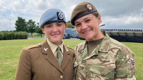 A teenage girl and her mum stand outside, smiling at the camera. They are both wearing military uniform, although the teenager's clothing is more formal, reflecting her involvement in a military parade.