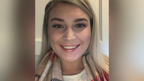 Close up of Leonie Wilkinson who has blond hair, brown eyes and brown eyebrows wearing a cream top and beige scarf with red, white and black patterns standing in front of a white cupboard. She is smiling.