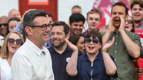 Andy Burnham is stood in front of a crowd of people who are cheering him. He is wearing a white shirt and glasses, and is smiling.