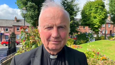 Bishop of Derry Donal McKeown has grey hair and is dressed in black. He is standing in front of flowers and trees in Brooke Park in Londonderryown