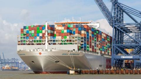 Cargo containers on ship. The containers are metal and of various colours and are piled high on the deck of a ship docked by a crane