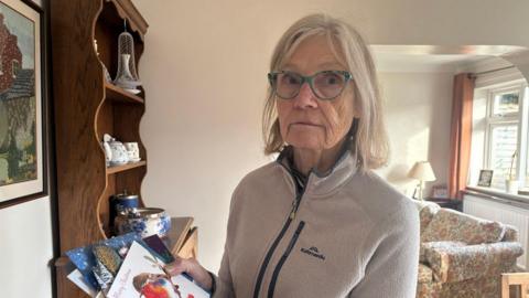 Catherine Simpson is in her living room wearing a fleece and holding Christmas cards. She is wearing glasses and looking unhappy. There is a dresser and a sofa in the background.
