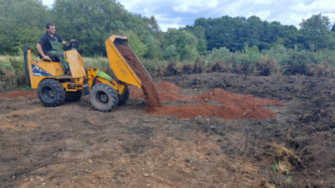 A man in a black shirt sits on a yellow digger as it unloads a red-coloured sand onto a large flat terrain of dirt. Trees can be seen in the distance.
