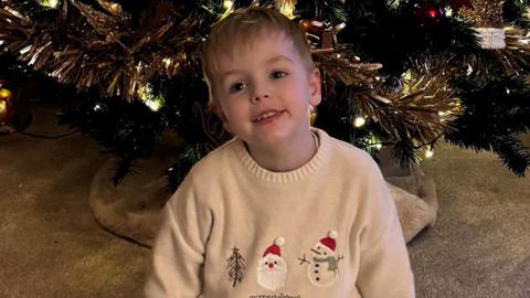 Freddie sitting smiling in front of a Christmas tree in a cream jumper with a Christmas tree, Santa and snowman on the front