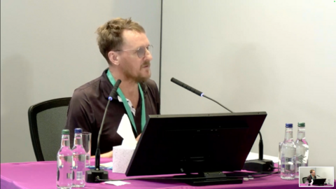 Stuart Ayris looks towards Baroness Lampard. He is wearing a black polo shirt and is sitting in front of a purple table with a laptop in front of him. He is wearing a green lanyard and has bottles of water in front of him. He is sitting in the inquiry room, with a microphone in front of him at Arundel House in central London
