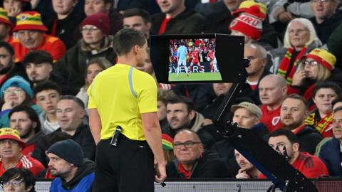 A referee in a yellow top stands in front of a football crowd while using VAR to make a decision