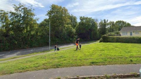 A general view of a green open space at the end of a road. An adult and child are walking across the grass. A person is on the pavement pushing a pushchair. Trees and a house are in the background.