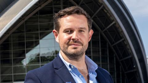 Sam Spittle - a man with short dark hair, stubble and a moustache, smiling while standing outside Glasgow Armadillo venue. He is wearing a dark blue jacket with light blue shirt.