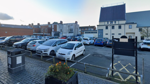 A wide shot of Playhouse car park in Whitley Bay. Cars are parked on a paved area with buildings in the background. At the front of the shot are raised flower beds with yellow and orange flowers.