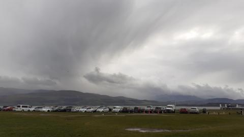 Grey skies overlooking a bay with mountains in the background and a row of vehicles in front of it