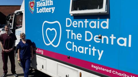 A man and a woman wearing dental tunics are standing in front of a large white van with a blue sign which reads: Dentaid The Dental Charity. 