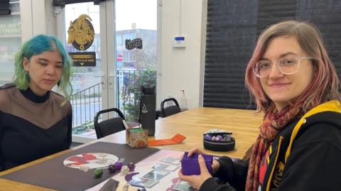 Evie and Giselle sit at a table facing and smiling at the camera. They sit opposite each other ready to play a board game. Evie has blue and green hair while Giselle has pink hair. A board game with cards and dice sits in between them on the table.