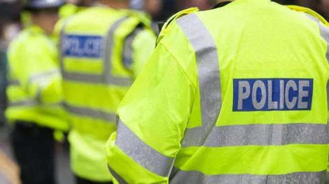 Three police officers standing outside with their backs turned to the camera. They are all wearing yellow fluorescent jackets with silver stripes along the arms and backs. On the back of each jacket there is a blue box with silver writing that says, POLICE.