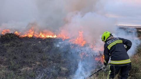 A picture of someone in a black uniform, with hi-visibility stripes on. They are beating a fire.