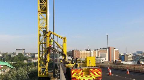 A highway maintenance vehicle parked on the Redheugh Bridge between Newcastle and Gateshead. The bright yellow vehicle holds a large piece of equipment which is stretched over the side of the bridge.  The equipment holds a large ladder which is standing vertical off the side of the bridge. A man in a hi-vis jacket is climbing on to the ladder.