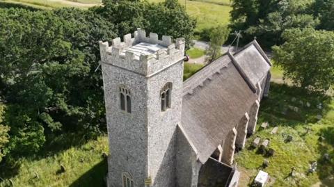 A church pictured from above, with a graveyard around and headstones. There are trees.