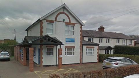 Rowlands Gill Medical Centre. The building is a white converted house with some red bricks on its corners. Two cars are parked in a small car park outside.