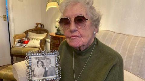 Win Crew holding a photo of her and Reginald when they were younger. She is wearing glasses, a dark green jumper, and necklace, and is sitting on a sofa.