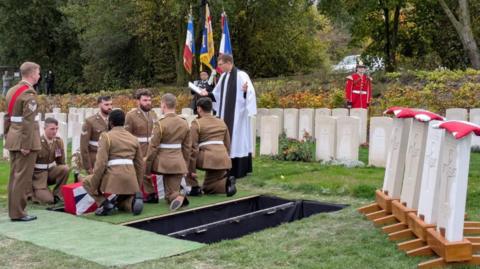 Serving soldiers, wearing full dress uniform, lower a Union flag-draped coffin into the ground, watched by a military padre.