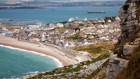 A shingle beach with a sea wall with a town built behind it. The photo is taken from up the cliff, looking down on the town. The town is on an island and the sea can be seen in front of and behind it.