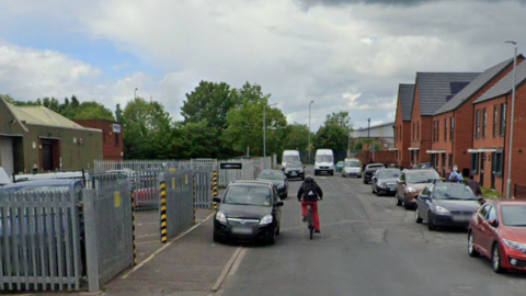 A residential road with houses along the right hand side and several cars parked. The left hand side of the road has an industrial building with trees along the back of it, and a cyclist pedalling away from the camera. 