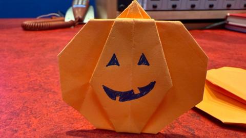 A origami orange pumpkin with a smile drawn on sits on a red desk