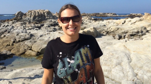 A woman wearing a black t-shirt with a colourful graphic is wearing sunglasses and smiling while standing on rocks at a beach