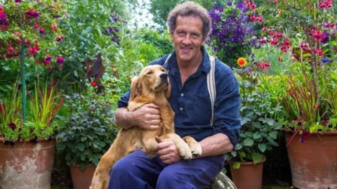 Monty Don, a middle-aged man with short dark curly hair, sits in a garden surrounded by large pots filled with plants and flowers. He is holding a golden retriever dog, which has its front paws on his lap.