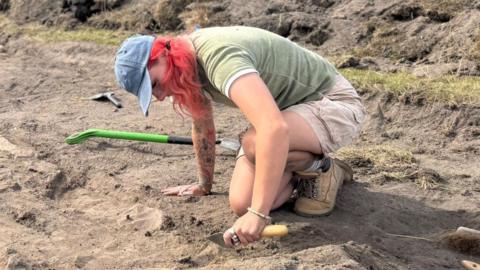 Meg Russell wearing a blue cap, green shirt, beige shorts, and work boots kneels in an archaeological excavation trench, carefully using a hand trowel to uncover features in sandy soil. Digging tools and spoil heaps surround the excavation area.