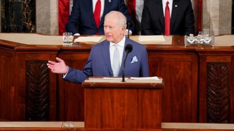 King Charles wears a blue striped suit with a while shirt and light blue tie. He is standing at a podium delivering a speech to Congress at the US House Chamber in Washington. He is gesturing with one hand out to his side