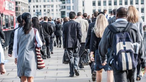 Lots of commuters walking along a pavement with their backs to the camera. Office buildings can be seen in the background