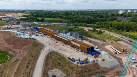 An aerial shot of a construction site next to a motorway, with green spaces and trees on the other side of the carriageway. A long brown structure is being built a few dozen feet from the motorway.