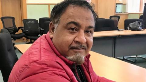 Faraz Ahmad wearing a red padded coat, sitting at a desk at the Nelson Plan for Neighbourhoods Board meeting. He has dark hair and a greying goatee beard.
