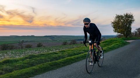 A cyclist cycles along a quiet country road at sunset. They're wearing a helmet and protective eyewear.
