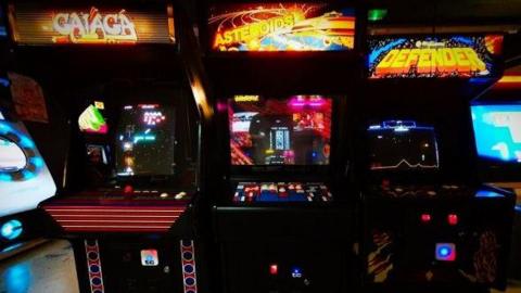 Three arcade machines with lit-up signs on the front and the screens are on. The room is dark and the lights are bright. 