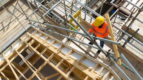 A bird's eye view of a builder wearing a hard hat and high visibility jacket works among trusses and scaffolding at a housing development.