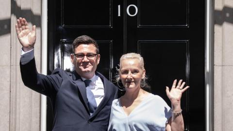 New Prime Minister Andy Burnham with his wife Marie-France van Heel outside 10 Downing Street, London. Andy Burnham wears a suit and a blue tie and Marie-France van Heel wears a blue dress. They are both waving.